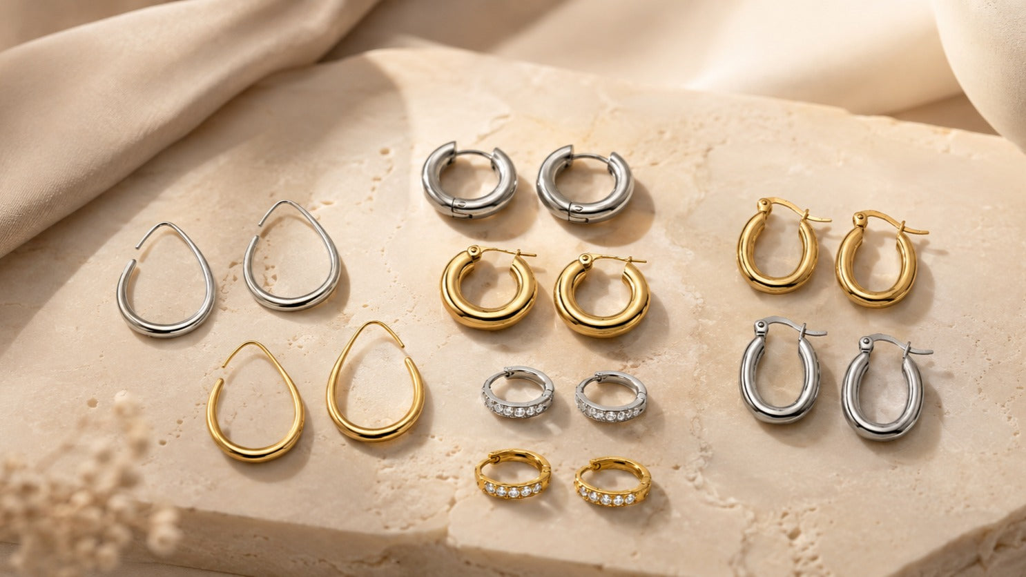 An assortment of gold and silver hoop, teardrop, and ring earrings arranged on a textured beige stone slab with draped fabric in the background.