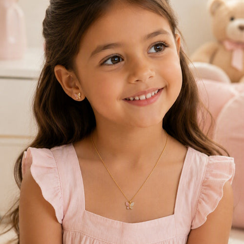 A young girl with long brown hair smiles while wearing a delicate gold butterfly necklace and a small matching stud earring, paired with a light pink ruffle-sleeve top. A soft-focus teddy bear is visible in the background.