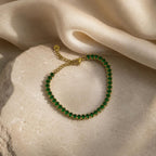 A gold chain bracelet adorned with a single row of green gemstones, displayed on a textured beige stone with draped cream satin fabric in the background.