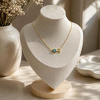 A delicate gold necklace with a small turquoise-blue stone tulip pendant is displayed on a cream-colored jewelry bust. The bust sits on a light wood base on a table, with a ceramic vase of dried flowers and soft sunlight in the background.