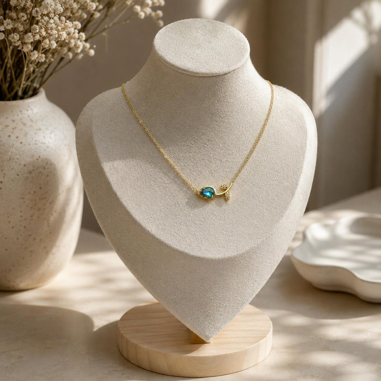 A delicate gold necklace with a small turquoise-blue stone tulip pendant is displayed on a cream-colored jewelry bust. The bust sits on a light wood base on a table, with a ceramic vase of dried flowers and soft sunlight in the background.