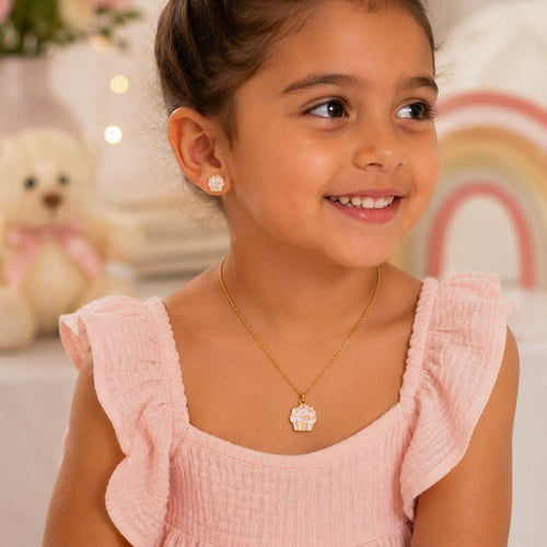 A young girl with her hair in a bun smiles while wearing a delicate gold necklace with a white cupcake-shaped pendant and a matching stud earring. She is wearing a light pink ruffle-sleeve top, with a soft, blurred background featuring a teddy bear and a rainbow.